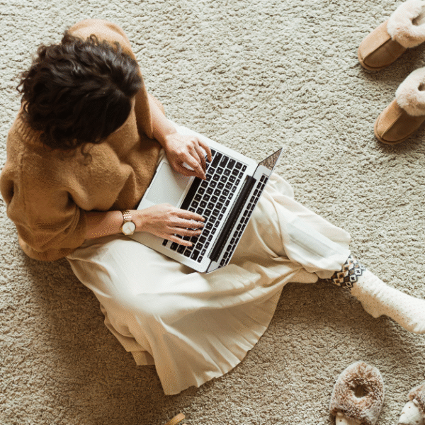 a woman sitting on a cosy rug on the floor, typing on her laptop